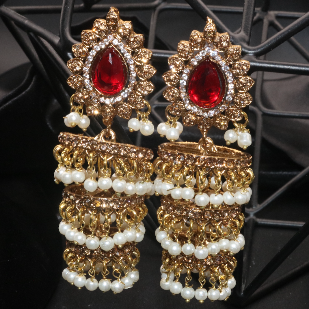 A pair of gold-colored jhumka earrings with white pearl-like beads and red gemstone accents, displayed against a black background.