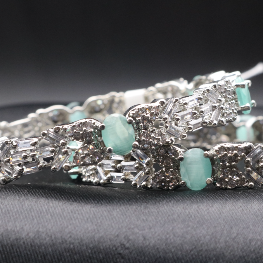 closer view of stainless steel bangles adorned with sparkling American diamonds, and teal gemstones displayed against a dark background.