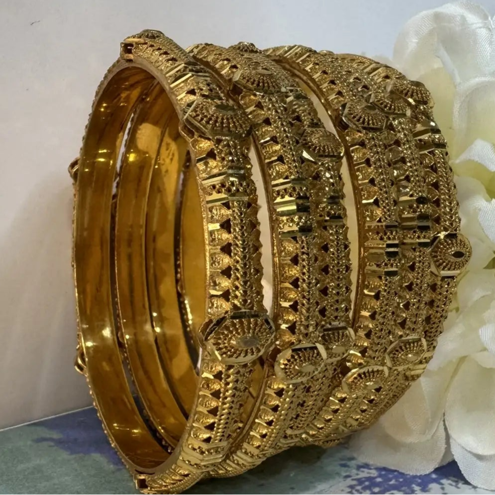 Gold bangles with intricate designs on a white surface with a white flower.