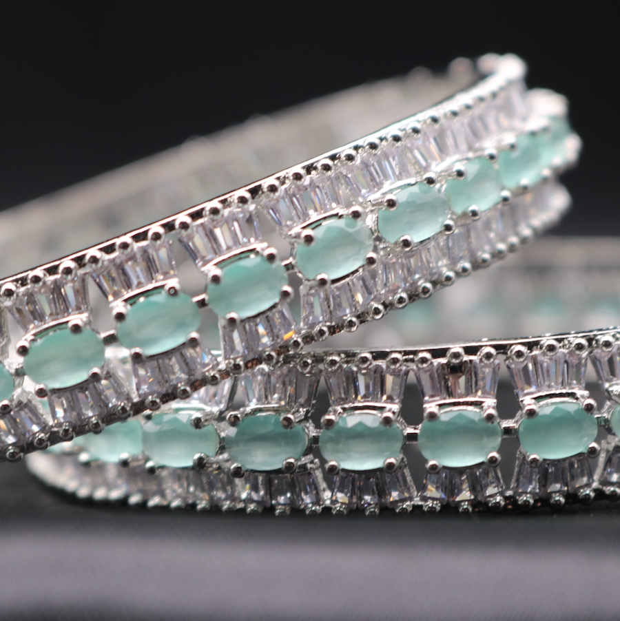 A set of stainless steel bangles adorned with sparkling American diamonds and teal gemstones, displayed against a dark background.