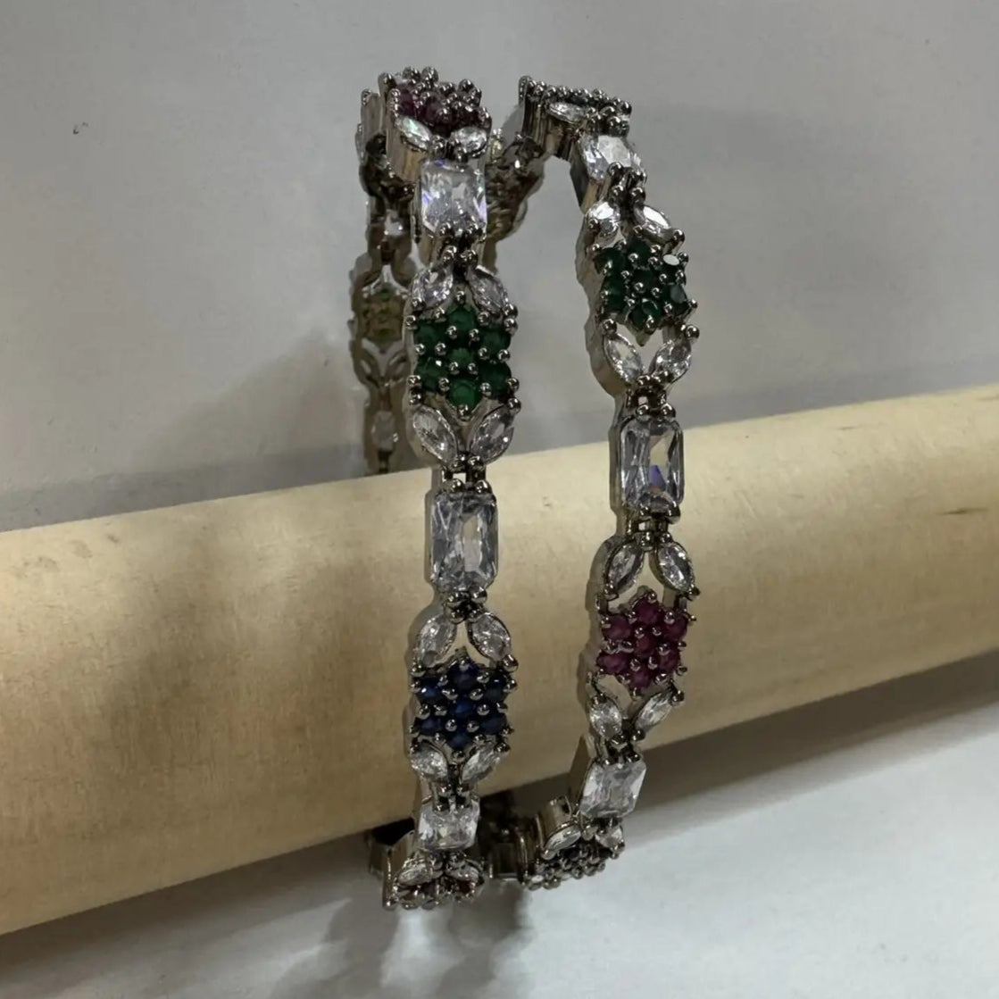 A multicolored diamond-encrusted bangles with vibrant hues of blue, green, and pink, set on a silver base, displayed on wooden stand against a neutral background