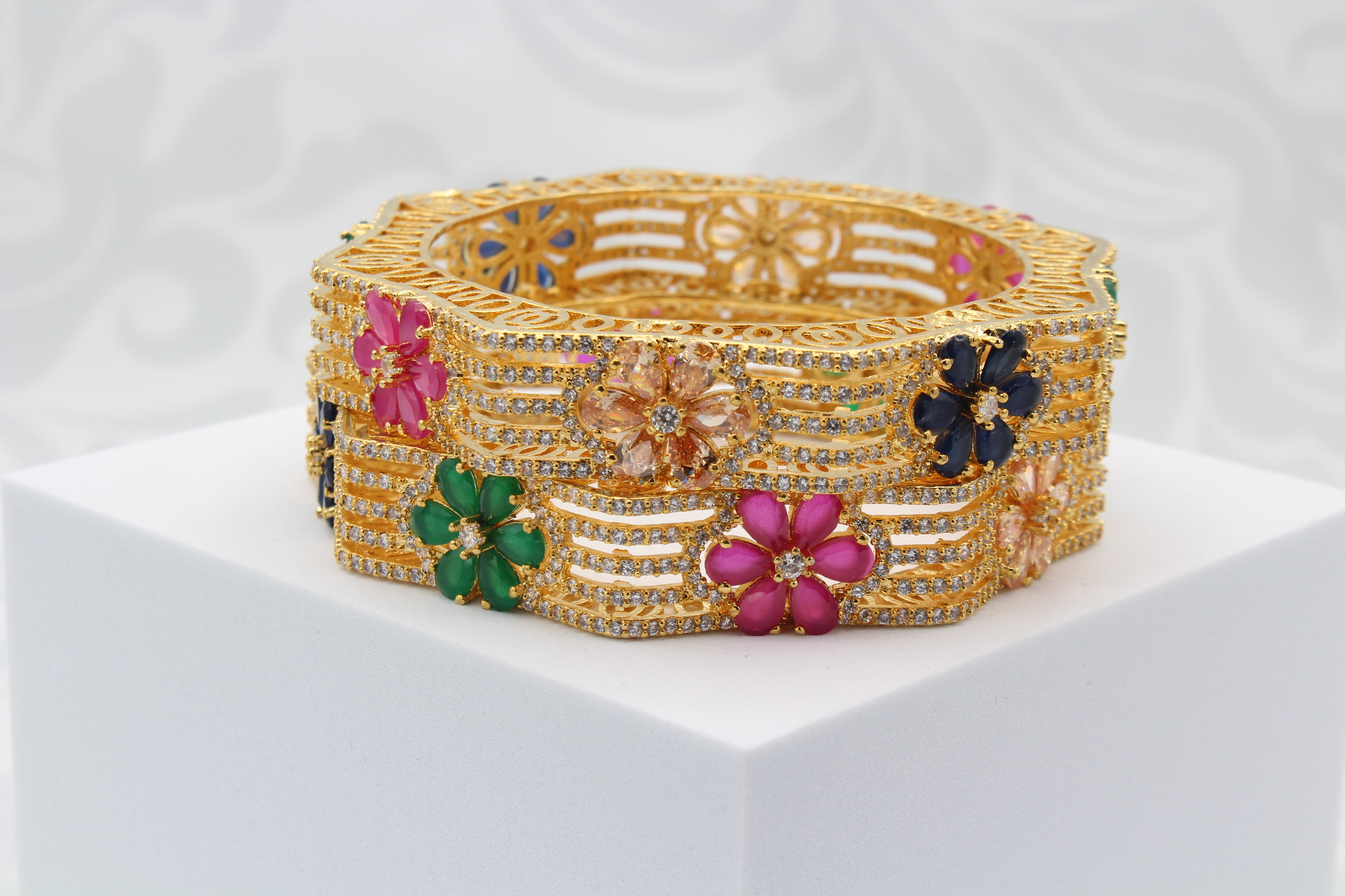 stacked gold-plated bangles with multicolor gemstones in a hexagonal design on white surface