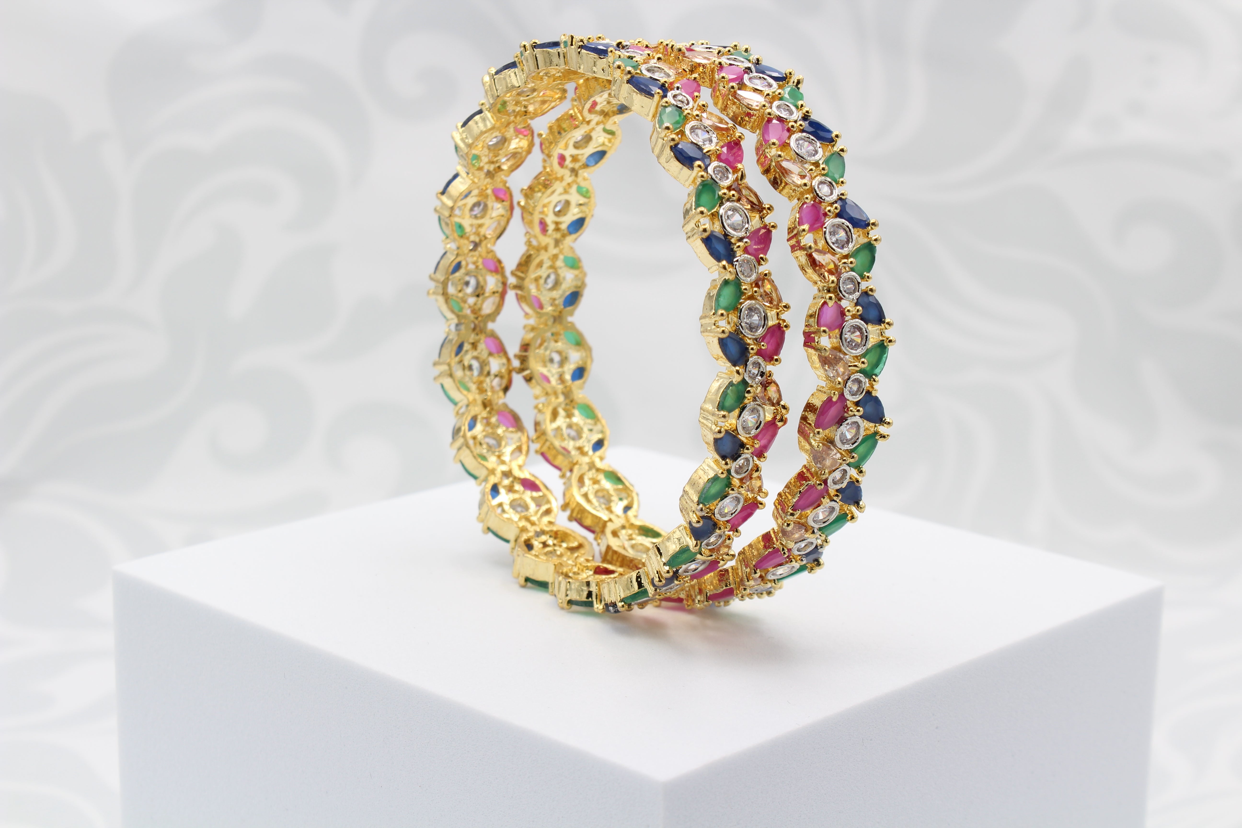 A pair of gold-toned leaf-shaped bangles adorned with multicolored stones displayed on a white platform with a patterned background.