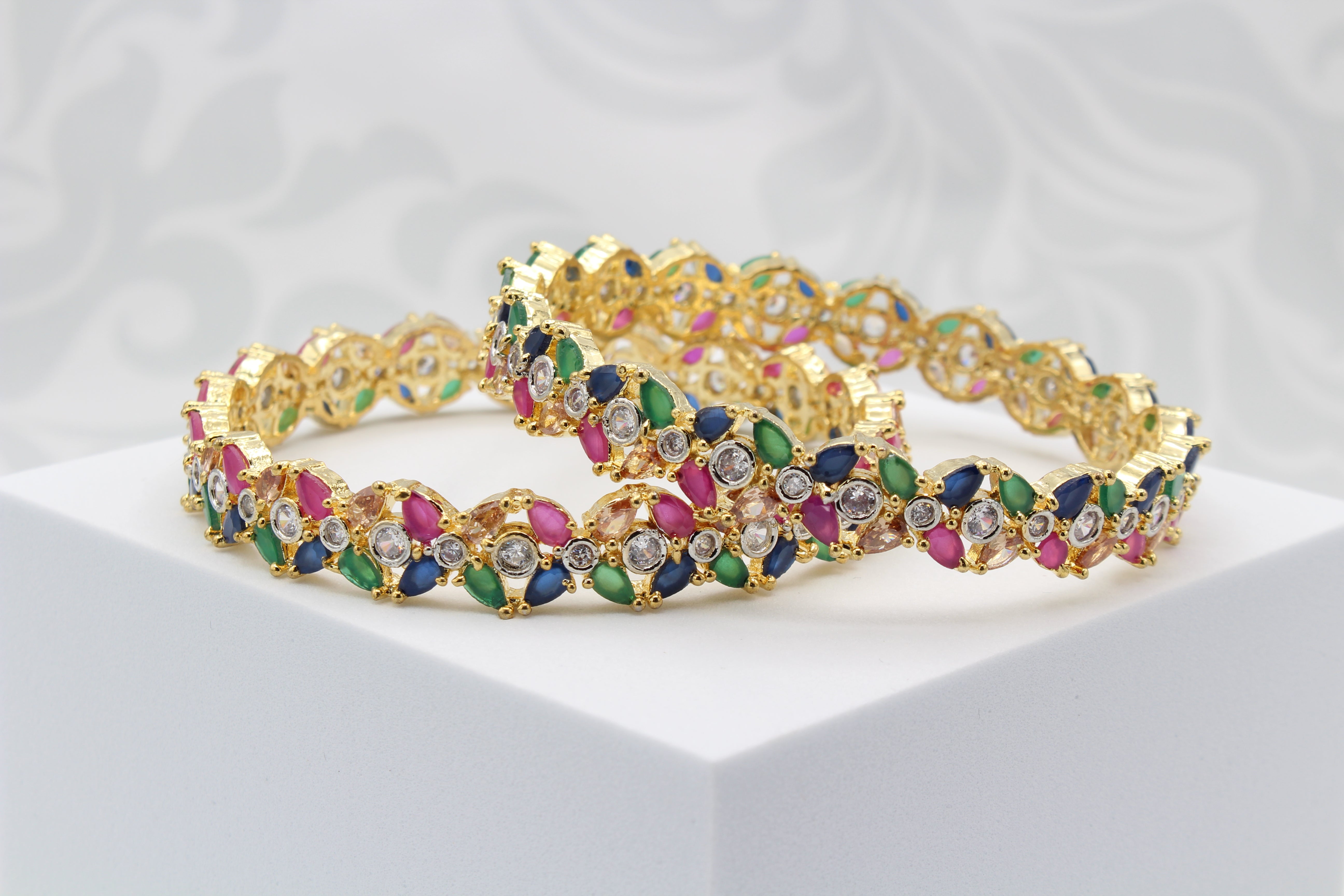 A pair of gold-toned leaf-shaped bangles adorned with multicolored stones displayed flat on a white platform with a patterned background.