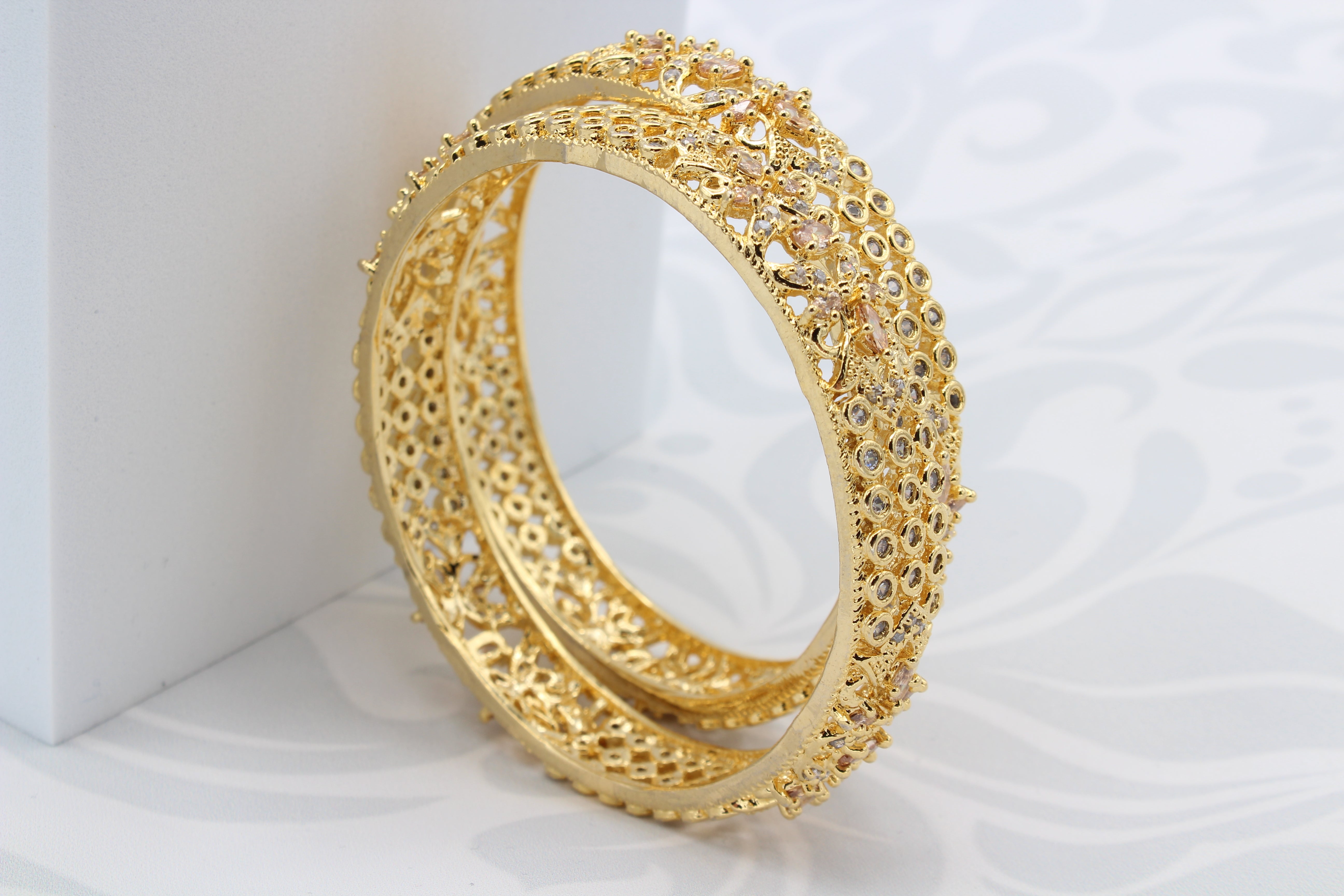 upright view of Gold plated pair of bangles with shimmering shine and intricate design on a white surface