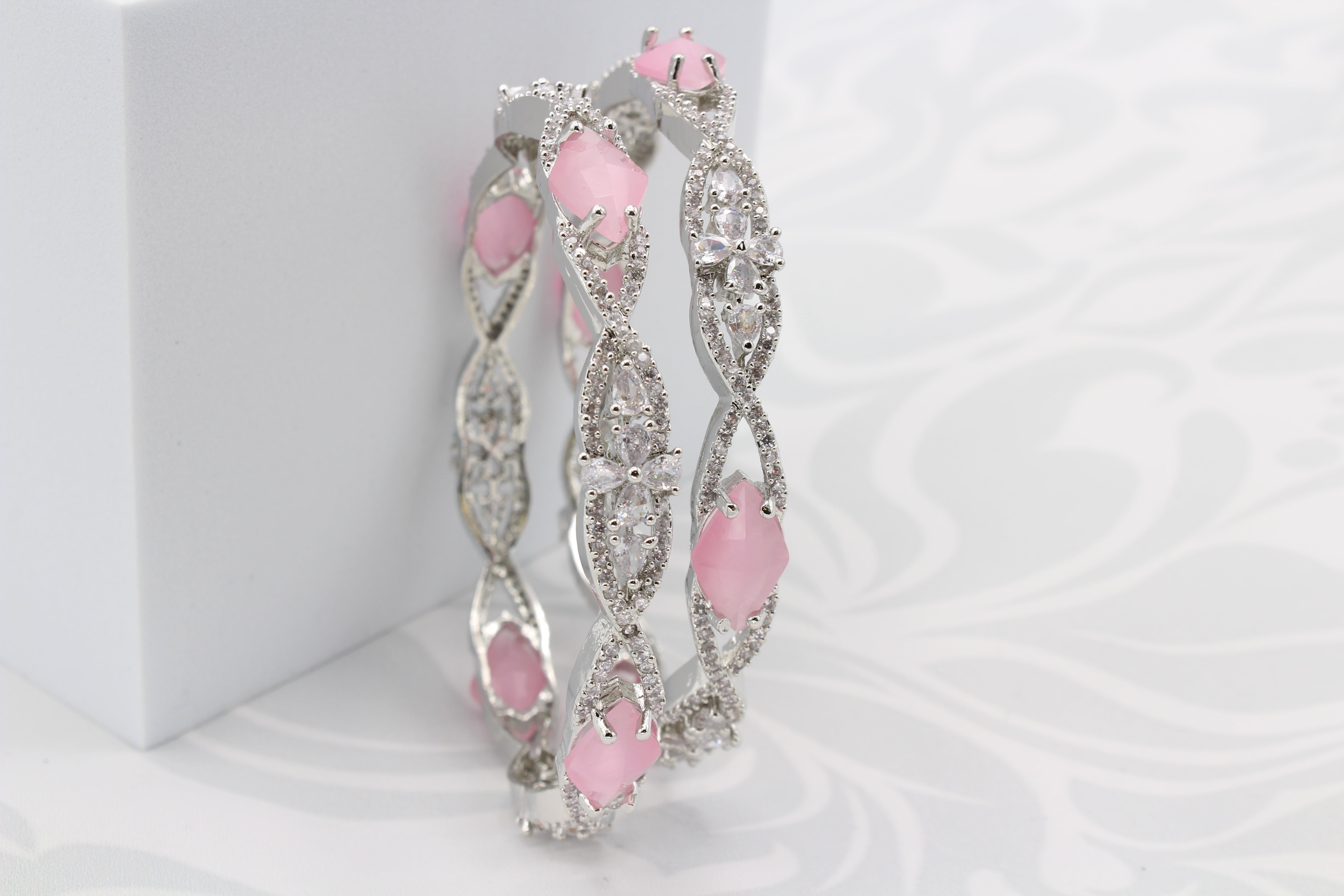 A pair of silver bangles adorned with pink rhinestones and American Diamond encrusted on a silver base, displayed on a white surface in an upright position